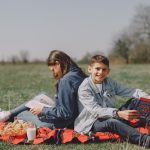 Smiling young boy in casual clothes sitting together with girl and reading interesting story while leaning back to back on checkered blanket on grass in park