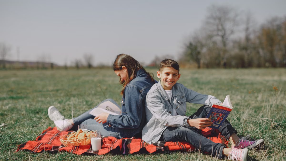 Smiling young boy in casual clothes sitting together with girl and reading interesting story while leaning back to back on checkered blanket on grass in park